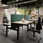 Modern office workspace setup with a light wood desk, dark green hexagonal office partition, black swivel office chairs, concrete floor, brick wall, and large windows looking out to greenery.