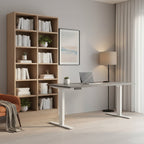 A modern height-adjustable standing desk with a gray top and white T-style legs, shown in a bright home office setting with a laptop, lamp, and framed wall art.
