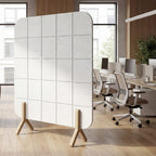 A modern white panel room divider with a grid pattern and wooden legs, shown in a bright, contemporary office lounge setting.