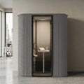 A modern gray privacy pod phone booth with a glass door, set against a minimalist beige wall in an office hallway.