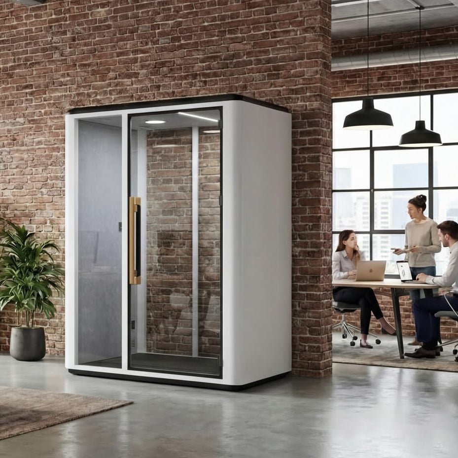 A modern office interior with a large white and glass privacy booth or soundproof room near a brick wall, alongside a wooden desk, office chairs, hanging lights, and people working in the background.